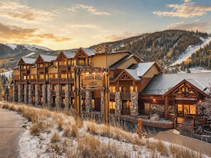 A lodge with multiple wooden buildings, a sign reading 'Hidden River Lodge,' and a mountainous backdrop. - Slopeside | Hot Tub | Garage | Walk to Lifts! (Keystone)