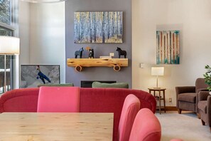 Ein modernes Wohnzimmer mit einem roten Sofa, einem Couchtisch aus Holz und einem Kamin. An der Wand hängen zwei Gemälde, ein Holzregal mit Dekorationen und eine Lampe auf einem Beistelltisch.