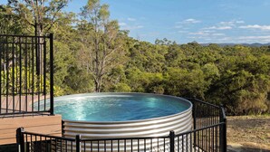 Een hottub omgeven door een houten terras en een metalen reling, met uitzicht op een bebost landschap en bergen op de achtergrond.