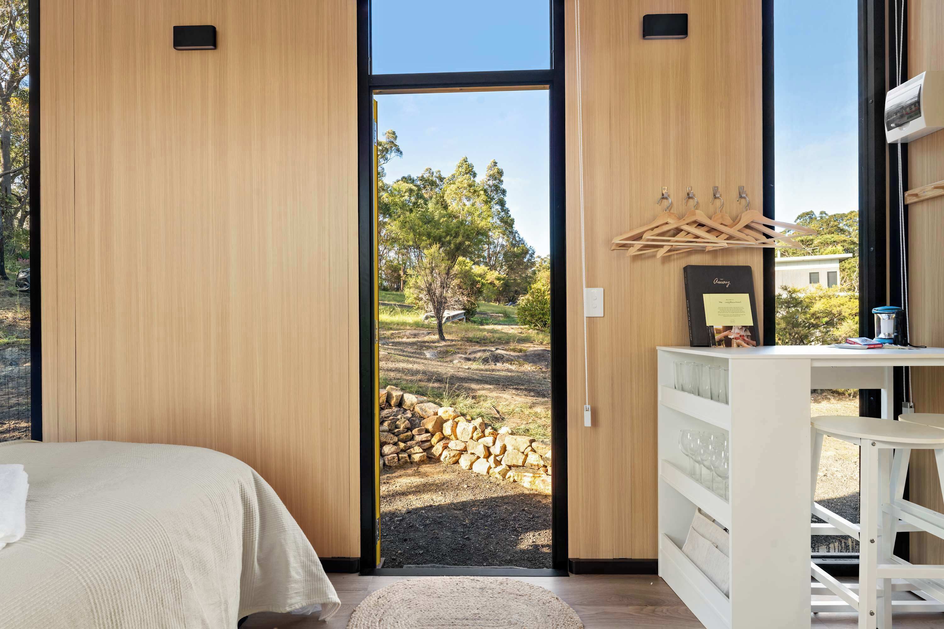 Un dormitorio con una cama, una puerta de madera que da a un espacio exterior con un montón de leños, y un aparador blanco con un espejo y un perchero.