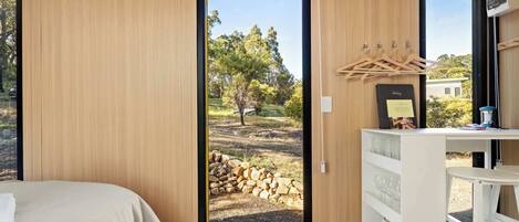 Un dormitorio con una cama, una puerta de madera que da a un espacio exterior con un montón de leños, y un aparador blanco con un espejo y un perchero.