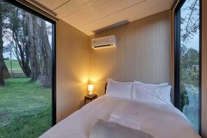 A bedroom with a bed, a nightstand with a lamp, an air conditioning unit, and a large window with a view of trees and grass.