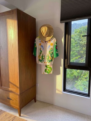 Un armario, una ventana con vistas a la vegetación y una persona con una camisa y un sombrero estampados.