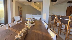Un vaste espace dédié à la salle à manger, avec une longue table en bois, des chaises en osier et un escalier moderne en arrière-plan.