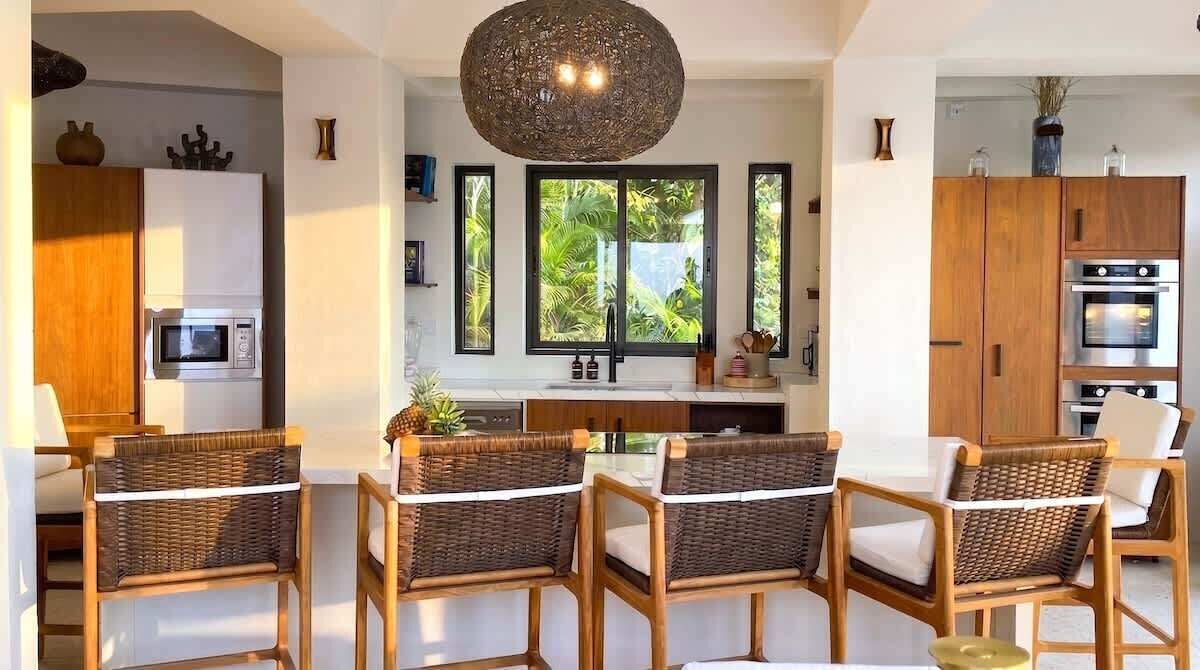 A modern kitchen with a central island, wooden cabinets, and a large window with a view of greenery.