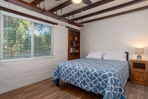 A bedroom with a bed, a window with blinds, a wooden bookshelf, and a nightstand with a lamp. - 1945 Ranch House / Tanque Verde / 2 BR 1 BA (Tucson)