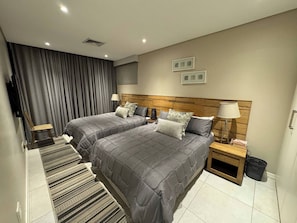A hotel room with two beds, a wooden headboard, a small bedside table with a lamp, a chair, a striped rug, and a large window with curtains. - Luxury 3 Bedroom Penthouse - Umhlanga Rocks (uMhlanga)