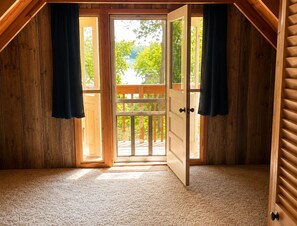 Una habitación con paredes de madera, piso alfombrado y un amplio ventanal que da a una terraza exterior con vistas a árboles y un lago.