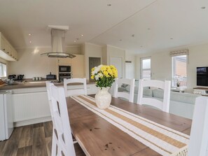 Una cocina moderna con armarios blancos, un comedor con mesa de madera y sillas blancas, y una sala de estar con sofá y televisor.