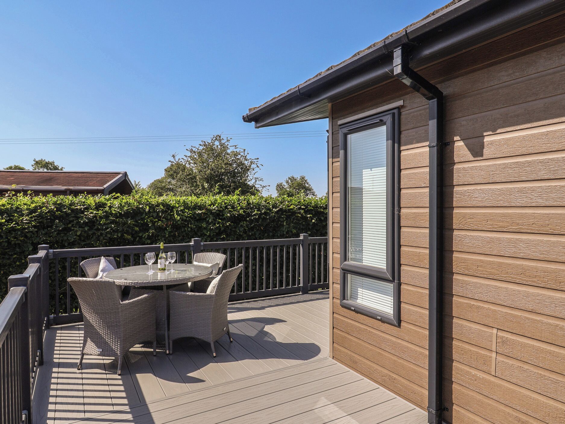 A wooden deck with a table and chairs, a window, and a hedge in the background.