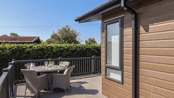 A wooden deck with a table and chairs, a window, and a hedge in the background.