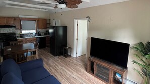A living room with a blue sofa, a flat-screen TV, a ceiling fan, and a kitchen in the background. - Terrific Scituate Rental ~ 8 min walk to sandhill beach (Scituate)