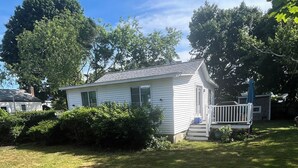 A small white house with a porch, surrounded by a well-maintained lawn and trees. - Terrific Scituate Rental ~ 8 min walk to sandhill beach (Scituate)
