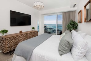 Ein modernes Schlafzimmer mit einem großen Bett, einem an der Wand montierten Fernseher, einem Holzkonsolentisch und Blick aufs Meer durch die Glastür.