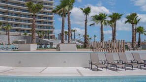 A hotel pool area with lounge chairs, palm trees, and a modern building in the background. - Beachfront Paradise 4br/4ba Elegant Condo With Breathtaking Views (Puerto Peñasco)