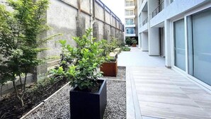 A narrow outdoor corridor with a concrete wall on one side and a building on the other. There are potted plants and a wooden bench along the walkway. - Rbk Nvo Estudio pasos del Movistar arena (Santiago)
