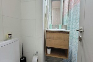 Une salle de bain avec un mur recouvert de carreaux blancs, un meuble-lavabo en bois avec un miroir, un porte-rouleau de papier toilette et un balai.