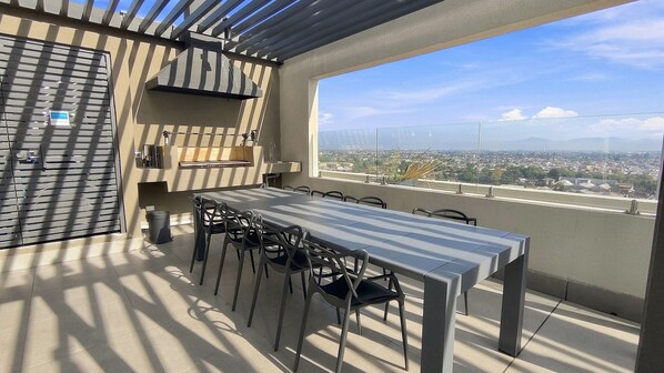 An outdoor balcony with a dining table and chairs, a grill, and a view of a cityscape.