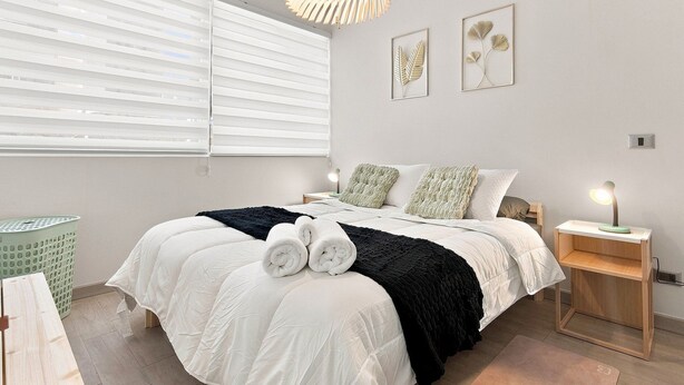 Een slaapkamer met een bed, kussens, een nachtkastje, een lamp en een raam met jaloezieën.