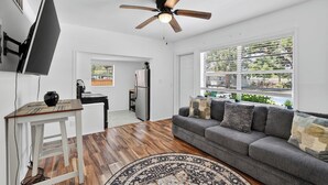 A living room with a sofa, a television, a ceiling fan, and a view of the outdoors through large windows. - Cozy 1-bedroom house in lovely Tampa with AC - 2706C (Tampa)