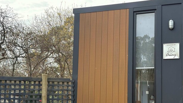 Un bâtiment moderne avec un revêtement extérieur en panneaux de bois, une porte en verre et une enseigne indiquant qu’il est géré par Tiny Away.