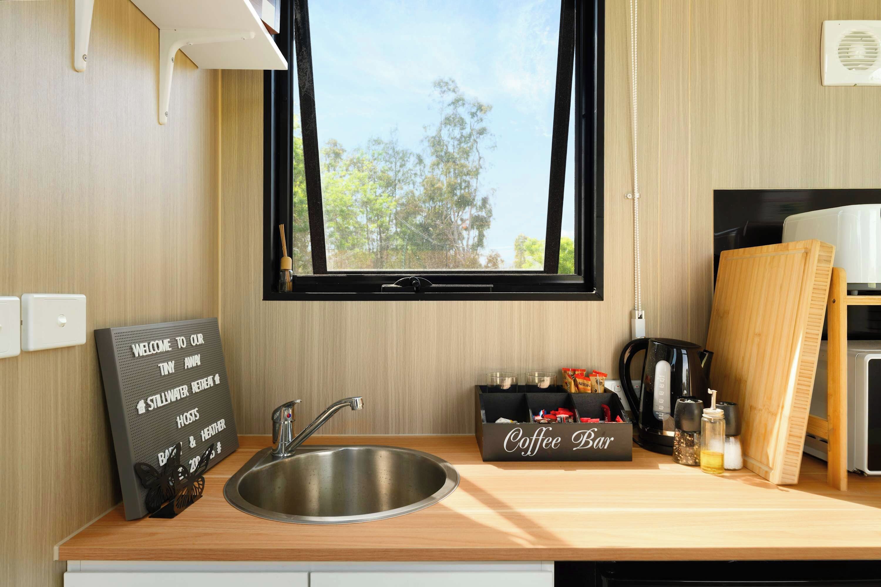 A kitchen with a stainless steel sink, a coffee bar sign, and a window with a view of trees.