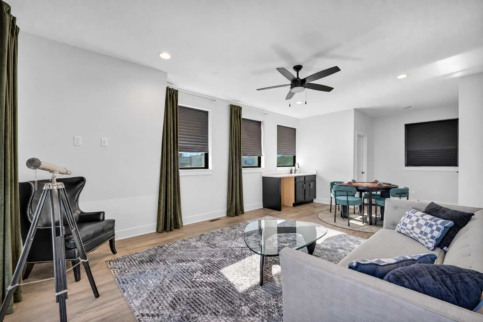 A modern living room with a glass coffee table, a sofa, a dining area with a table and chairs, a ceiling fan, and a telescope on a tripod.