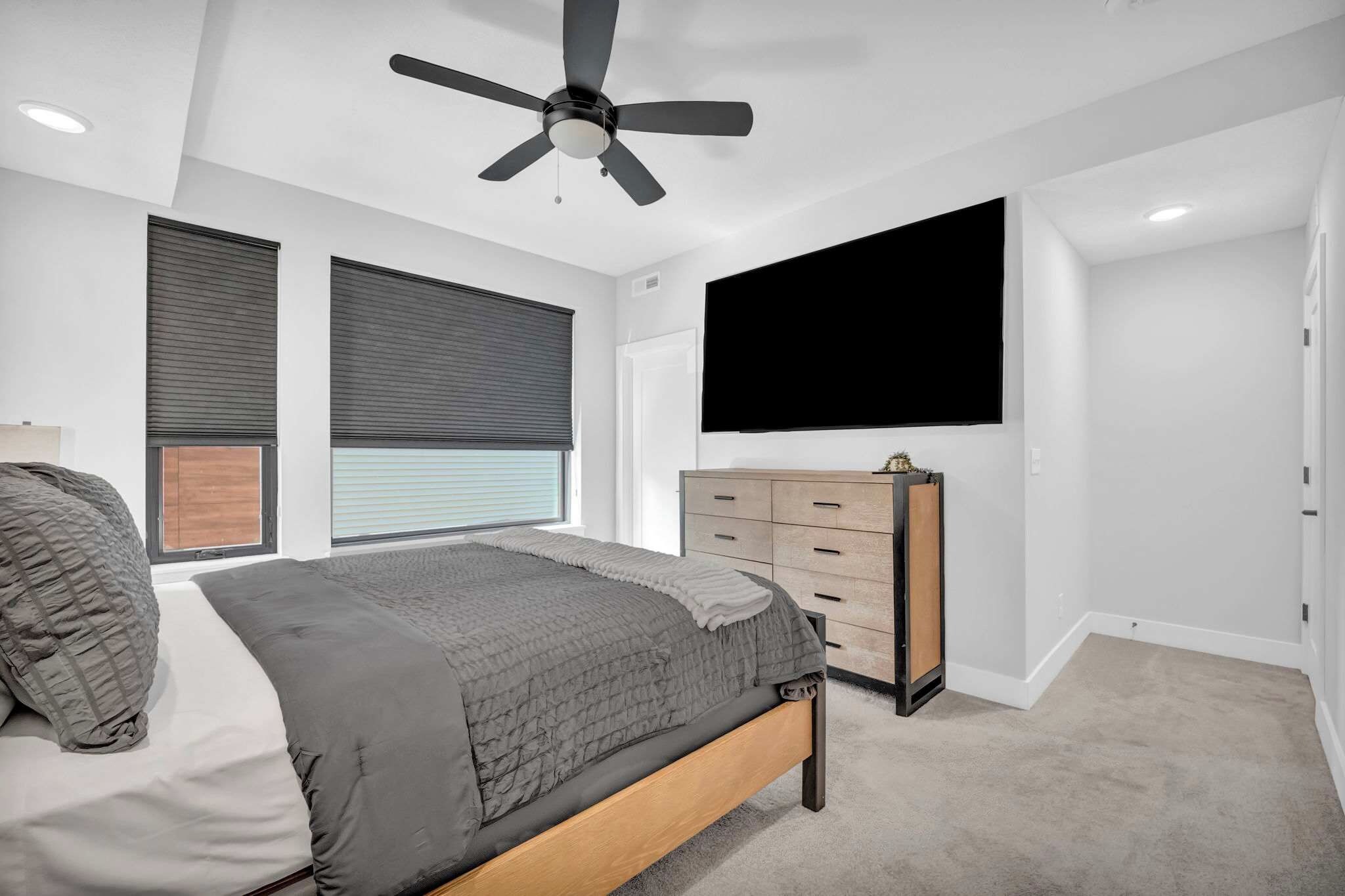 A bedroom with a bed, a dresser, a ceiling fan, a window with blinds, and a TV mounted on the wall.