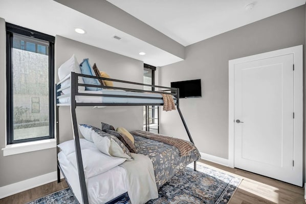 A bunk bed with a TV mounted on the wall above it, a door on the right, and a window on the left.