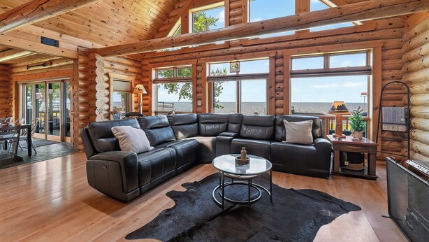 A log cabin interior with a leather sectional sofa, a glass coffee table, and a large window overlooking a body of water. - Gorgeous log home in Southern Door- Waterfront retreat (Luxemburg)