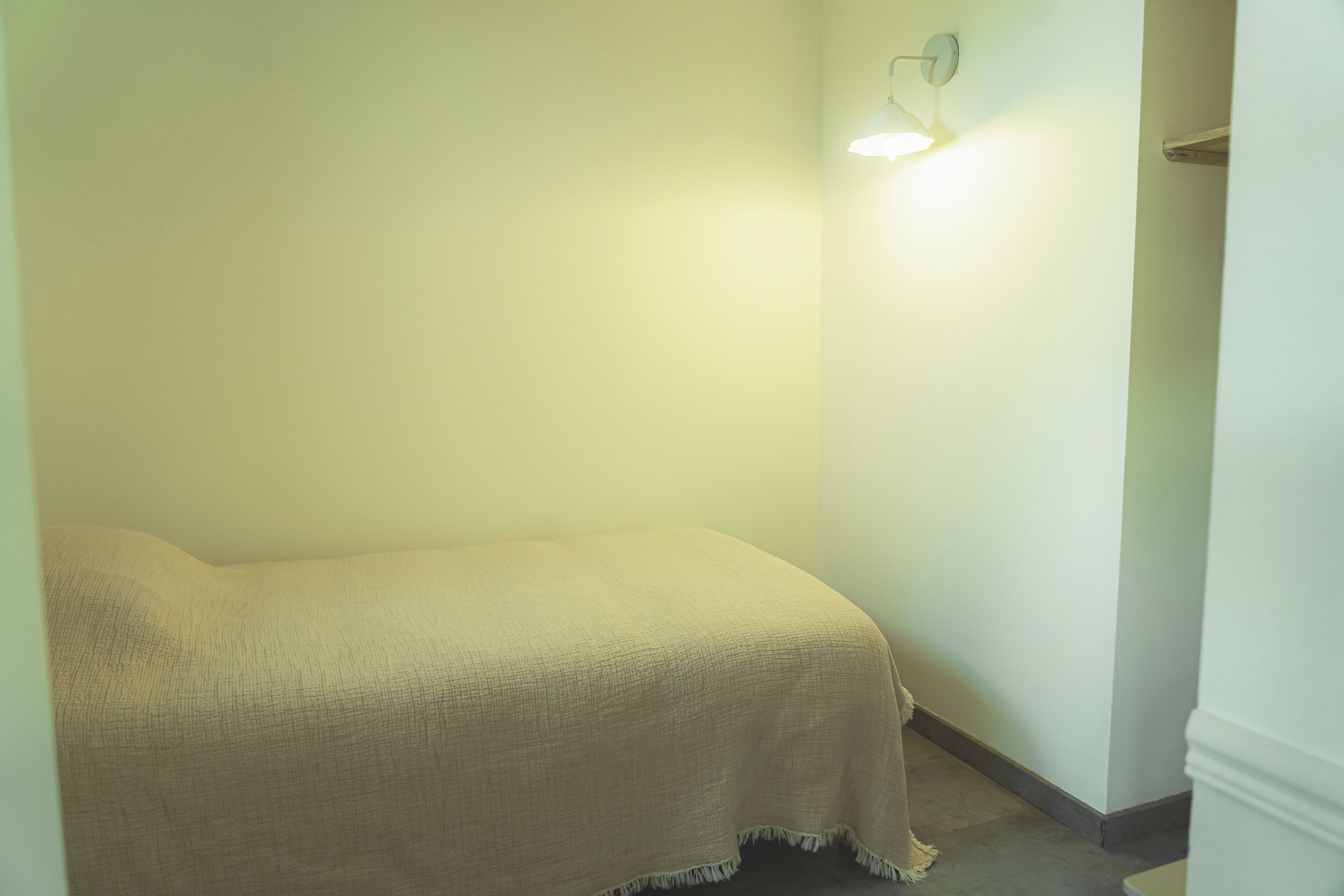 Ein kleiner, schwach beleuchteter Raum mit einem Einzelbett, das mit einer beige farbenen Decke bedeckt ist, einer Wandleuchte und einer Tür auf der rechten Seite.