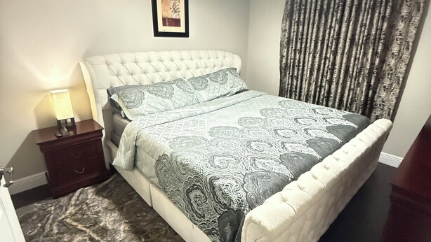 Un dormitorio con cabecera acolchada, colcha estampada, lámpara de noche, un cuadro en la pared y una ventana con cortinas.