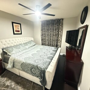 Un dormitorio con cama, ventilador de techo, televisor y ventana con cortinas.