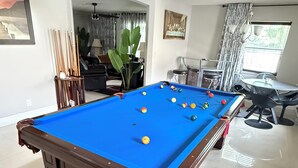 Una mesa de billar en una sala con comedor, una planta grande y un cuadro en la pared.