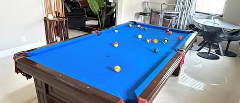 A pool table in a room with a dining area, a large plant, and a painting on the wall.