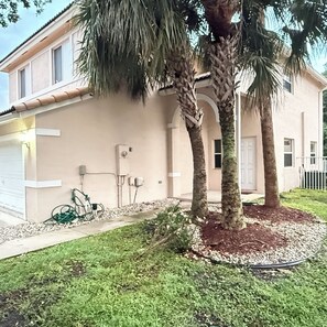 A residential building with a white exterior, a garage, and a well-maintained lawn with two palm trees. - Delightful 2-bedroom house with AC in Coconut Creek (Coconut Creek)