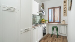 A compact kitchen with white cabinetry, a green stool, a window with curtains, and a 'KITCHEN' sign on the wall. - Concettina Apartment sea view for up to 5 people! (Massa Lubrense)