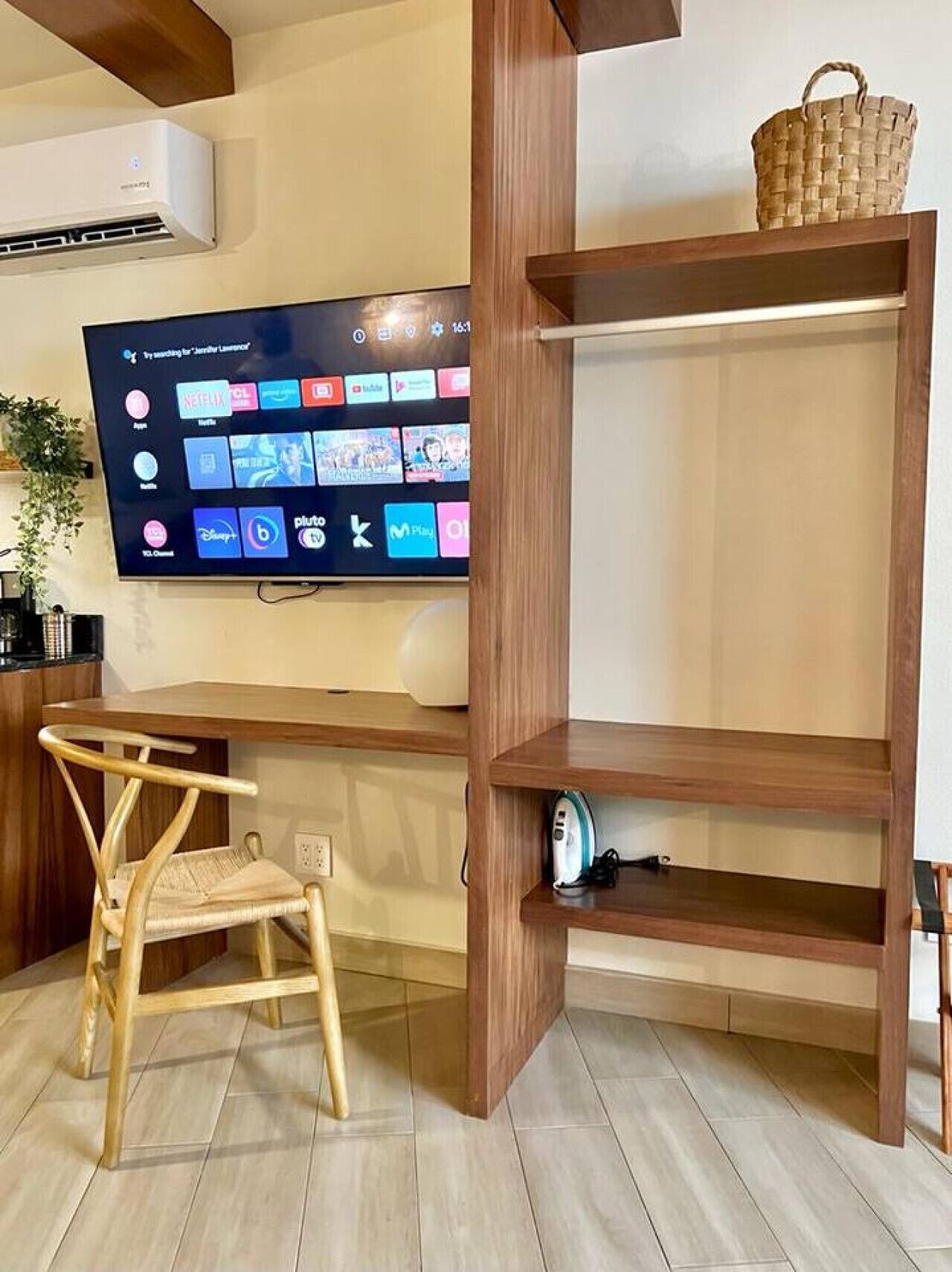 Una camera con una scrivania in legno, una televisione fissata al muro, un cesto in vimini sulla scrivania e una sedia in legno.