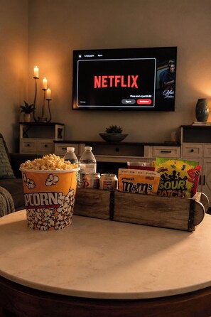 Ein Wohnzimmer mit einem Fernseher, auf dem das Netflix-Logo zu sehen ist, sowie Popcorn, Limonade und Snacks auf einem Tisch.