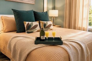 A neatly made bed with a quilted cover, two pillows, a bottle of champagne, and two glasses of orange juice on a tray.