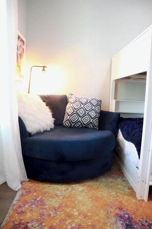 Ein Eckzimmer mit einem dunkelblauen Sofa, einem gemusterten Kissen, einer flauschigen weißen Decke, einem weißen Etagenbett und einem farbenfrohen Teppich.