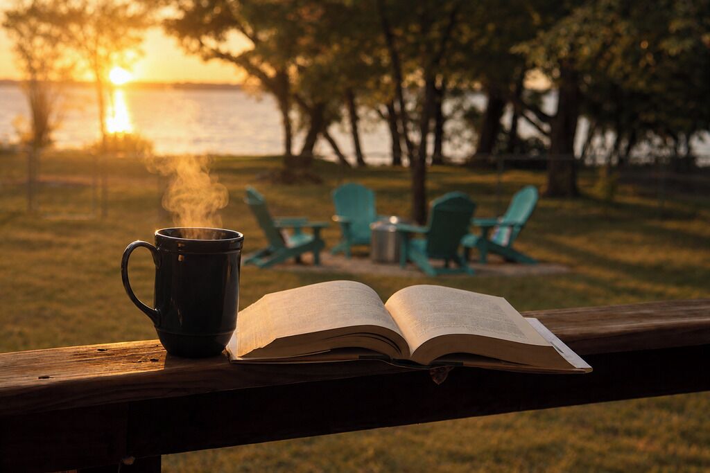 Un libro abierto y una taza humeante sobre una barandilla de madera, con vista a sillas y árboles al fondo.