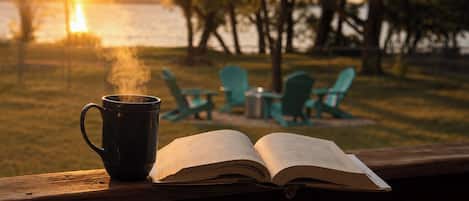 Un livre ouvert et une tasse fumante posés sur une rambarde en bois, avec en arrière-plan des chaises et des arbres.