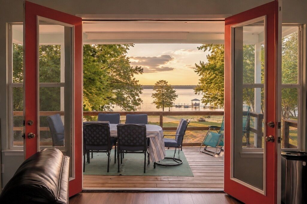 Porta aperta che dà su una terrazza con tavolo e sedie, con vista su un'area d'acqua e alberi.