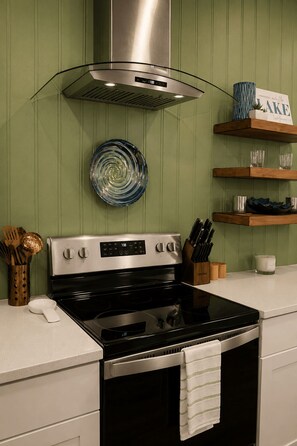 Una cocina con estufa de acero inoxidable, campana extractora y un plato en la pared. Hay estantes de madera con cristalería y un letrero que dice 'LAKE'.