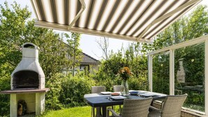 En udendørs patio med et stentabel, fletstole, grill og udsigt til et hus og træer.