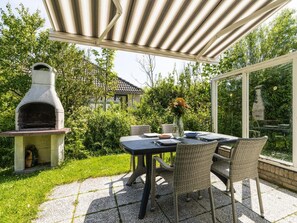 Eine Terrasse im Freien mit einem Steintisch, Korbstühlen, einem Grill und Blick auf ein Haus und Bäume.