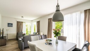 Et moderne hotelværelse med spiseplads, en sofasæt, et stort vindue med gardiner og udsigt til grønne omgivelser.