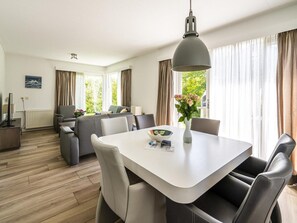 Ein modernes Hotelzimmer mit Essbereich, einer Sofagarnitur, einem großen Fenster mit Vorhängen und Blick auf Grünflächen.