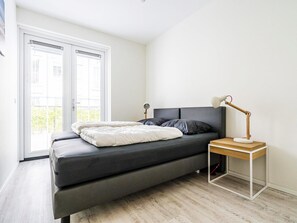 A bedroom with a bed, a bedside table with a lamp, and a view through a window. - Apartment Near Koudekerke Beach (Koudekerke)